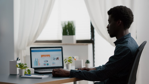 Gerente afro-americano trabalhando em casa, analisando gráficos com vendas e receitas, trabalhando remotamente no laptop da sala de estar. usuário de computador negro usando comunicação on-line na web para negócios na internet