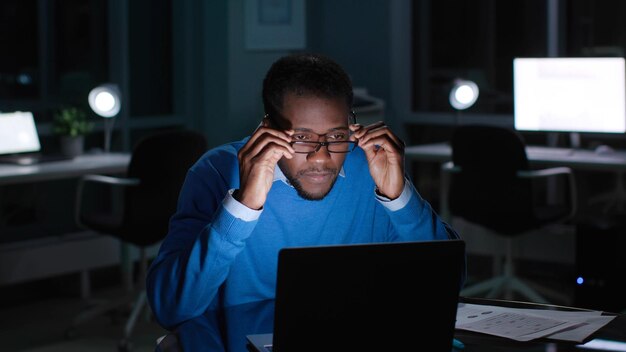 El gerente afroamericano con anteojos lee el correo electrónico en una computadora portátil que trabaja tarde en la noche