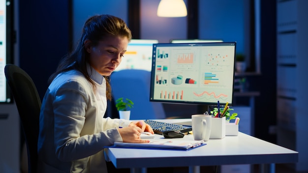 Gerente agotado tratando de terminar el proyecto empresarial respetando la fecha límite trabajando por la noche frente a la computadora tomando notas escribiendo en el cuaderno. Empleado cansado horas extras sentado en el escritorio en el lugar de trabajo