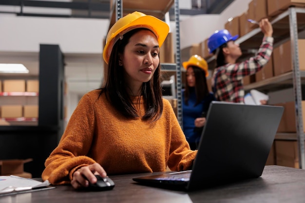 Foto gerente de almacén trabajando en la gestión automatizada de inventario en una computadora portátil mientras está sentado en el escritorio de la sala de almacenamiento. operador de almacén con casco amarillo analizando los datos de los pedidos de los clientes en la computadora