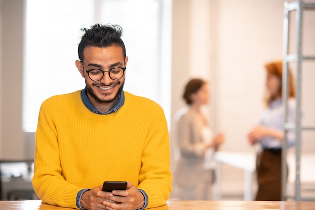 Gerente árabe jovem bonito e positivo de óculos, parado no balcão do escritório e enviando mensagens de texto sms no telefone