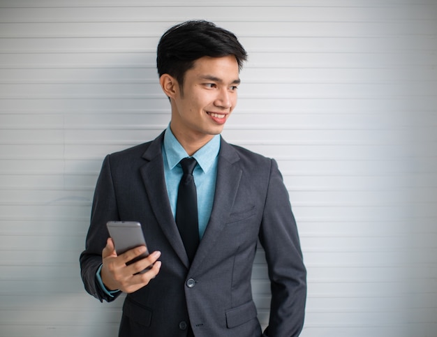 Gerente asiático joven feliz en traje sonriendo y navegando por teléfono móvil mientras está de pie contra la pared gris