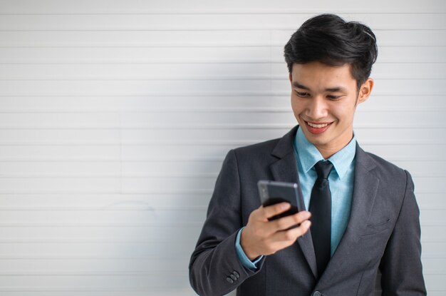 Gerente asiático joven feliz en traje sonriendo y navegando por teléfono móvil mientras está de pie pared gris