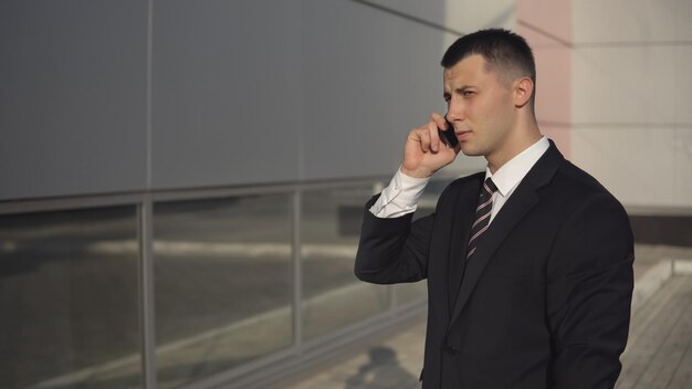 Gerente atractivo en traje formal hablando por teléfono mientras está de pie en el fondo del edificio de gran altura en un día soleado. copyspace