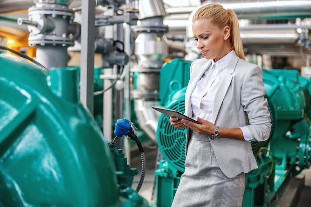 Gerente bem-sucedida e trabalhadora sorridente de terno em pé na planta de aquecimento com um tablet nas mãos e verificando a turbina