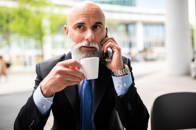 Gerente calvo y elegante con barba blanca tomando un café al aire libre mientras está al teléfono