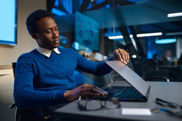 Gerente cansado fechando o laptop, estilo de vida do escritório à noite. homem à mesa, interior escuro do centro de negócios, local de trabalho moderno