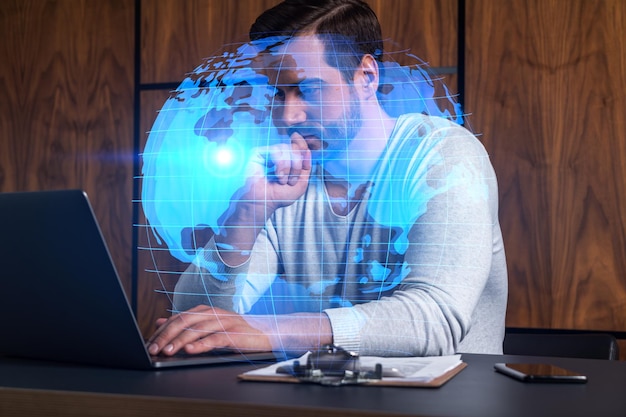 Un gerente caucásico serio en ropa casual usa una computadora portátil para desarrollar una nueva cadena de suministro mundial Globo de doble exposición y holograma de mapa