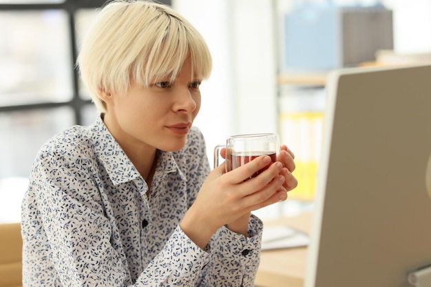 Gerente com caneca de vidro de chá olha para o monitor do computador em estudos de mulheres no escritório da empresa