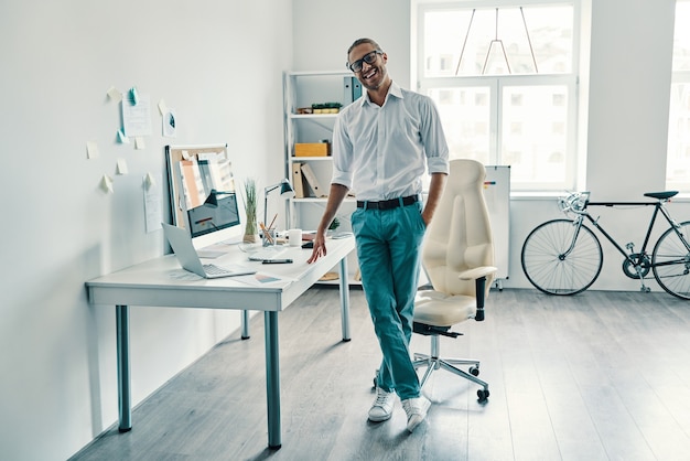 Gerente confiante. comprimento total de um jovem bonito em camisa e jeans sorrindo enquanto estava no escritório