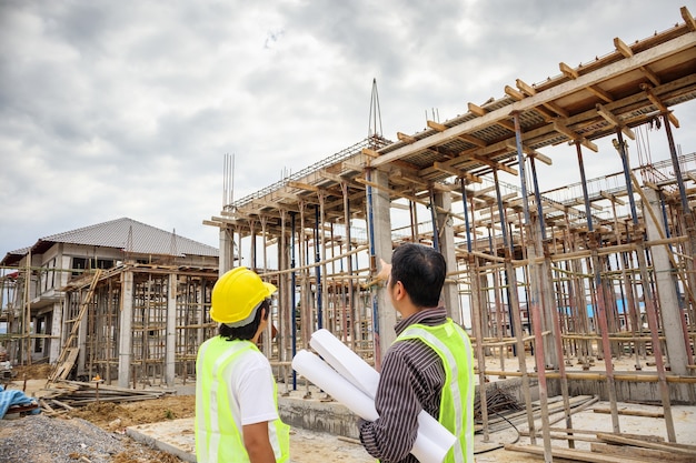 Gerente de construcción de hombre de negocios asiático y trabajador ingeniero en casco protector sostienen papel de planos a mano en el sitio de construcción de viviendas