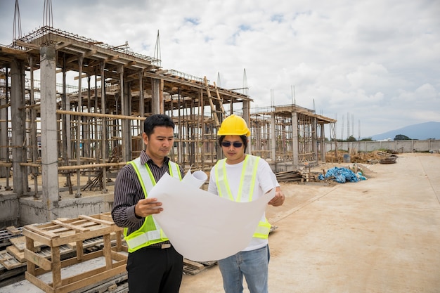 Gerente de construcción de hombre de negocios asiático y trabajador ingeniero en casco protector sostienen papel de planos a mano en el sitio de construcción de viviendas