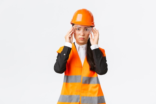 Gerente de construcción de mujer asiática joven de aspecto serio sigue el protocolo de seguridad, usa gafas protectoras y casco antes de ingresar a la empresa, de pie fondo blanco
