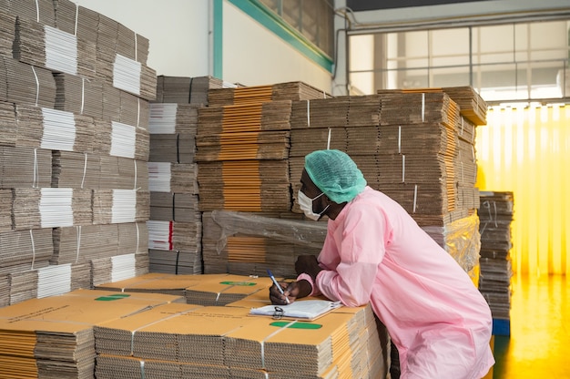 Gerente de control de inventario de hombre africano en ropa estéril comprobando el orden del paquete de caja de cartón apilado en el almacén en la planta de procesamiento de bebidas