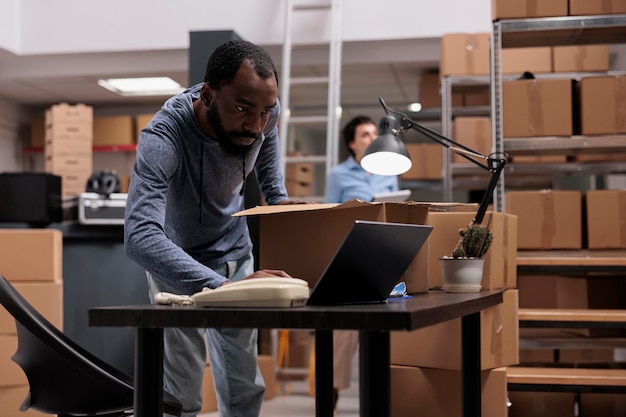 Foto gerente de armazém afro-americano verificando carga de estoque no computador laptop enquanto prepara pacotes, colocando pedidos de clientes em caixas de papelão. equipe trabalhando no departamento de entrega no armazém