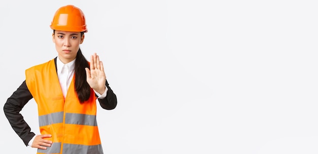 Gerente de construção de arquiteta asiática decepcionada com aparência séria na área de trabalho usando capacete de segurança mostrando gesto de parada proibir ação proibir invasão de fundo branco