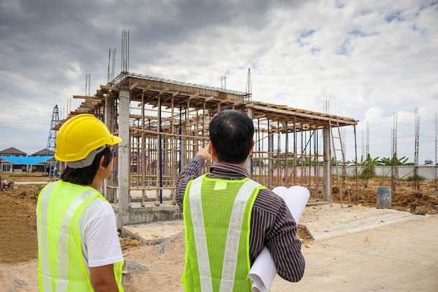 Gerente de construção de homem de negócios asiáticos e engenheiro trabalhador com capacete protetor segurando papel de planta disponível no local de construção da casa