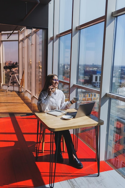 Gerente de empresária em uma camisa branca fazendo trabalho de escritório e sorrindo chefe feminina europeia bem sucedida em óculos ópticos para correção de visão posando em uma mesa em um espaço de trabalho