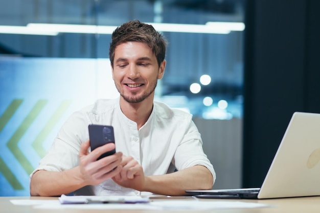 Gerente de empresário jovem sentado no escritório em sua mesa segurando um celular digitando mensagens fazendo ligações Funciona em laptops e documentos Foto aproximada