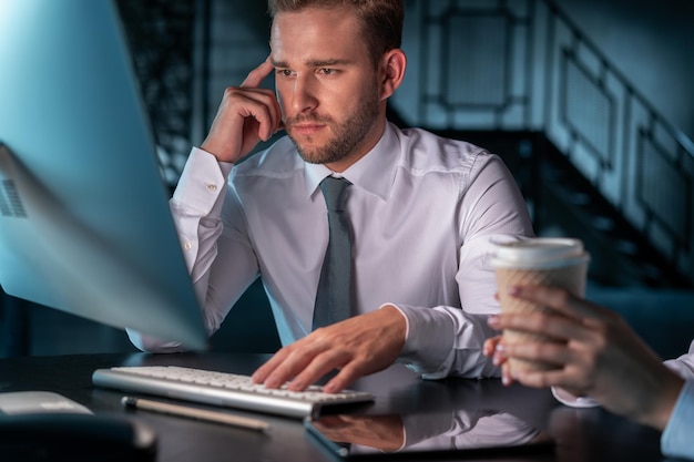 Gerente de escritório homem sério pensando na frente do monitor do computador mão no teclado Gerentes masculinos e femininos em camisas brancas trabalhando no escritório tarde da noite Conceito de trabalho em equipe