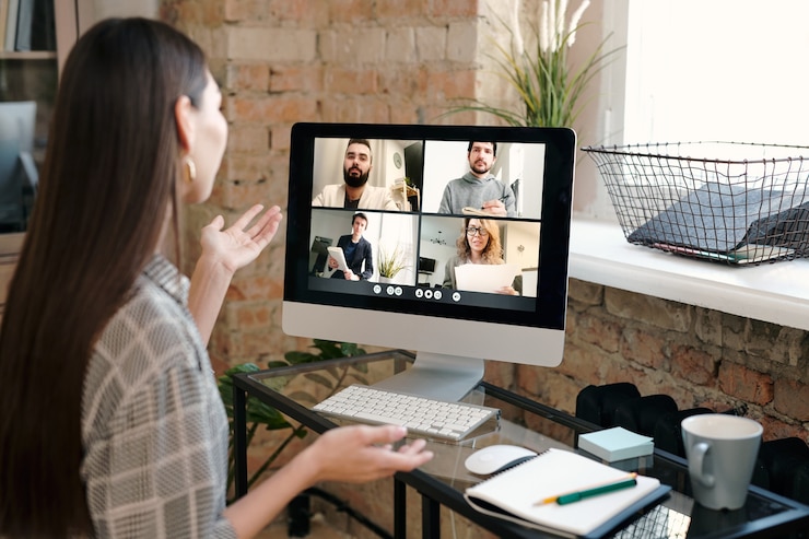 Foto gerente de escritório jovem contemporânea com longos cabelos escuros, sentada à mesa em frente ao monitor do computador e falando com as pessoas na tela