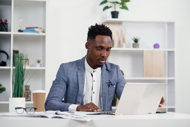 Gerente de escritório jovem de pele escura grave trabalha com diferentes relatórios e laptop de mesa em seu local de trabalho na sala de reuniões no moderno centro de negócios.