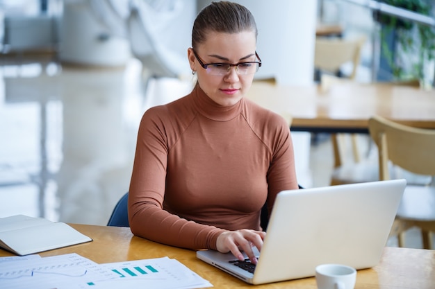 Gerente de garota bem-sucedida em seu próprio escritório com grandes janelas faz um novo plano de negócios de desenvolvimento econômico em um laptop com uma xícara de café. Mulher de negócios com documentos sentada a uma mesa de madeira