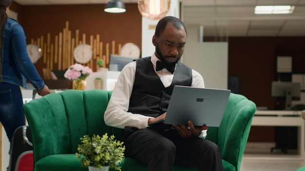 Gerente de hotel focado trabalhando em laptop sentado no lobby olhando para a tela e acompanhando as reservas para gerenciar reservas Bellboy em uniforme trabalha na indústria da hospitalidade Tiro portátil