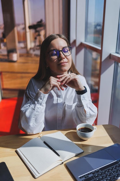 Gerente de mulher europeia senta-se em uma mesa enquanto trabalha em um laptop e bebe café em um café uma jovem sorridente usa óculos e uma camisa branca trabalho autônomo e remoto estilo de vida feminino moderno