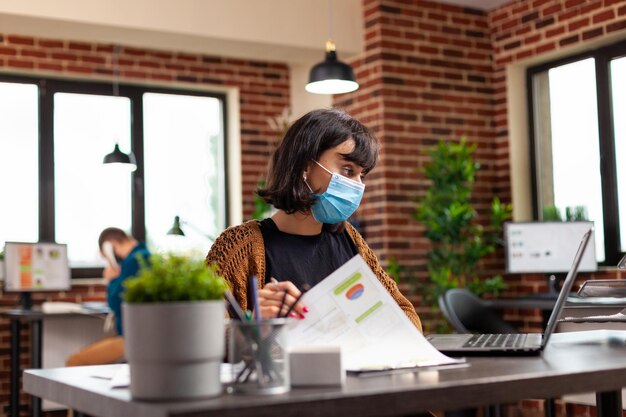 Foto gerente ejecutivo con mascarilla de protección médica para prevenir la infección con covid19 analizando gráficos comerciales escribiendo estrategia de gestión en computadora portátil. empresaria trabajando en ideas de proyectos