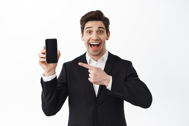 Gerente emocionado, oficinista apuntando a la pantalla del teléfono inteligente y sonriendo feliz, mostrando la aplicación o sitio web, demostración de la aplicación, pared blanca