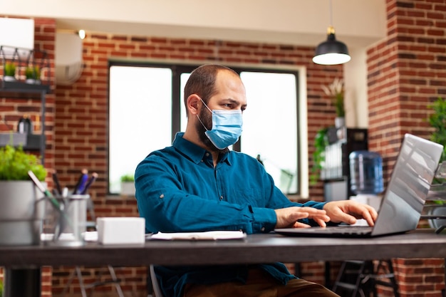 Foto gerente executivo com máscara facial de proteção para proteger a infecção com estratégia de marketing de digitação de coronavírus no teclado do laptop analisando o relatório de negócios. empresária trabalhando em estatísticas financeiras