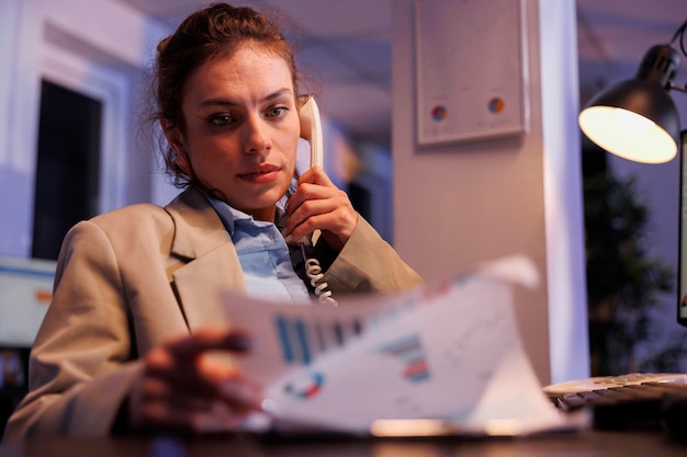 Gerente executivo falando no telefone fixo com funcionário remoto, analisando o relatório de gráficos de estratégia da empresa. Empresária trabalhando tarde da noite no escritório de inicialização. conceito corporativo