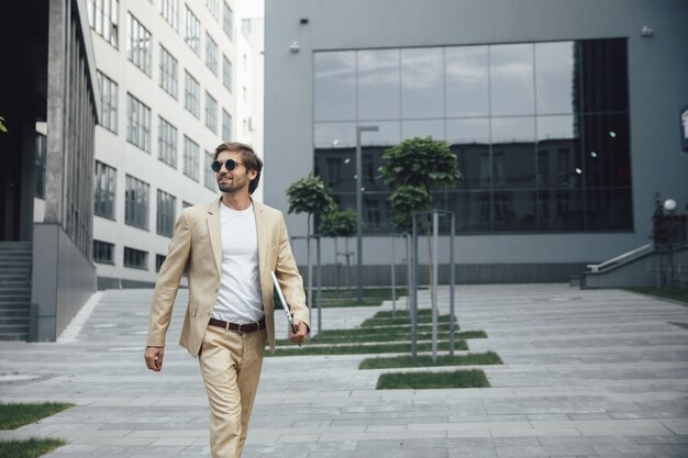 Gerente de éxito alegre en ropa formal caminando cerca del centro de negocios y llevando una computadora portátil inalámbrica