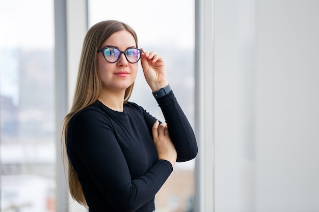 Una gerente exitosa en su propia oficina con grandes ventanales está de pie y observa la ciudad. Mujer de negocios con gafas en el fondo de grandes ventanales