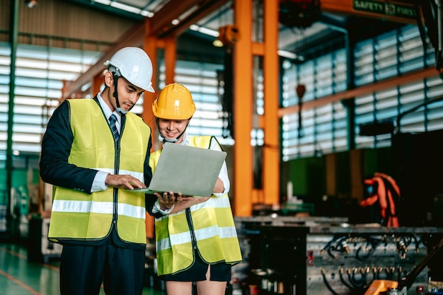 El gerente de fábrica y el trabajador con casco de seguridad usan una computadora portátil y una aplicación corporativa para discutir el resultado de la inspección Concepto de trabajo en equipo de la industria manufacturera