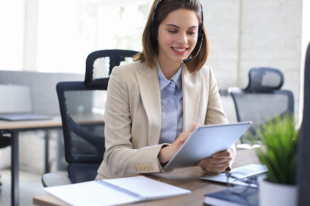 Gerente feminina alegre sentada na mesa do escritório e executando tarefas corporativas usando conexão sem fio em dispositivos digitais