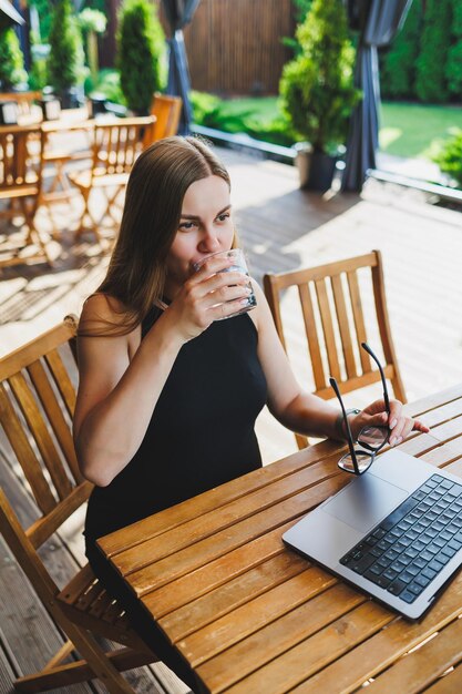 Gerente feminina freelancer desfrutando da pausa de café no terraço de verão de um café aconchegante trabalhando remotamente no laptop sorrindo olhando para o laptop Trabalhando online no verão