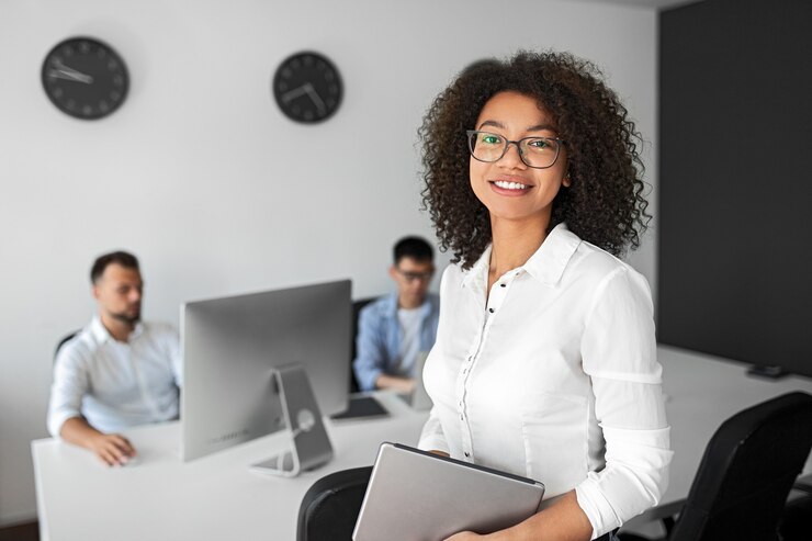 Foto gerente feminina moderna positiva em camisa branca formal e óculos, segurando o tablet e olhando para a câmera, em um escritório moderno trabalhando com colegas de computador