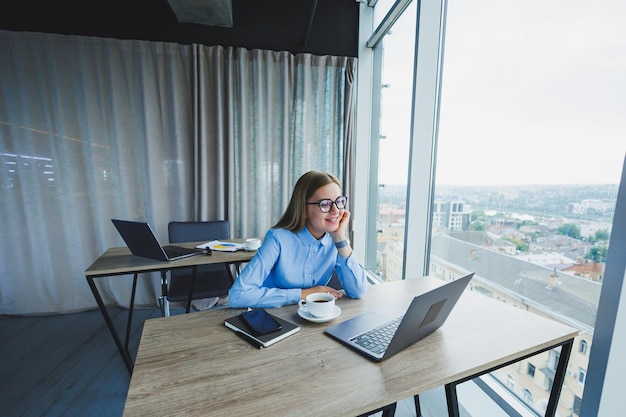 Gerente feminina sorridente adulta de óculos e camisa formal senta-se em uma mesa e olha para um laptop Escritório moderno espaçoso e brilhante com uma grande janela