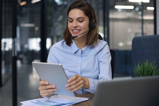 Gerente feminino alegre sentado na mesa do escritório e executando tarefas corporativas usando conexão sem fio em dispositivos digitais.