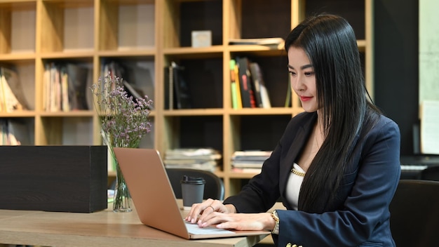 Gerente feminino asiático atraente sentado no escritório contemporâneo e usando computador portátil