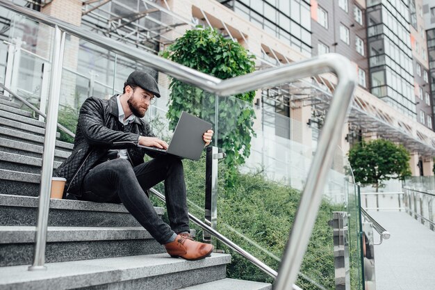 Gerente jovem e bonito bebendo café e trabalhando em um laptop enquanto está sentado ao ar livre na escada