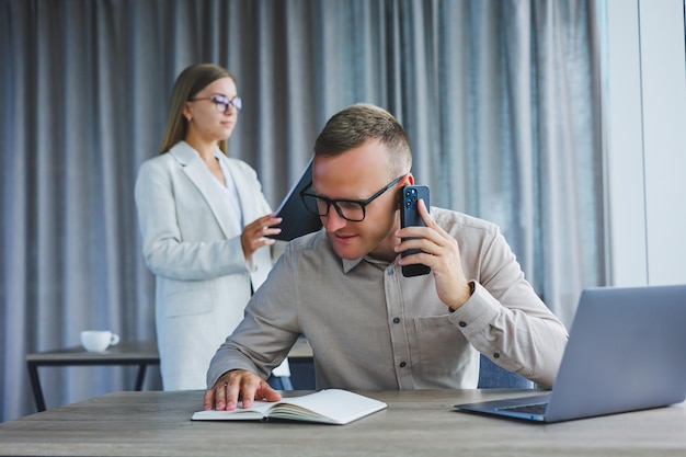 Un gerente masculino está hablando por un teléfono móvil mientras está sentado en una mesa con una computadora y un bloc de notas Ambiente de trabajo en una oficina con grandes ventanales