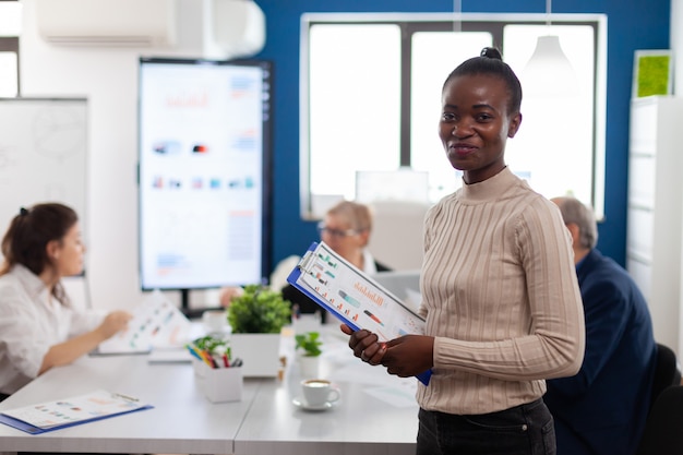 Gerente de mujer africana mirando a la cámara sonriendo, sosteniendo el portapapeles, mientras diversos compañeros de trabajo hablan en segundo plano. Gerente que trabaja en la puesta en marcha profesional de negocios financieros, lugar de trabajo de la empresa moderna