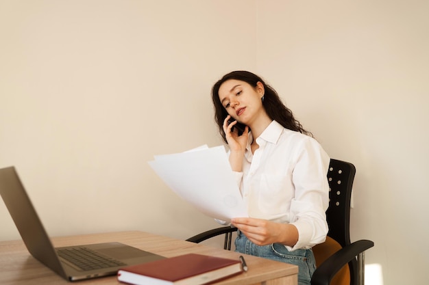 Gerente de mujer alegre hablando por teléfono haciendo papeleo y trabajando en la computadora portátil Chica jefa trabaja en la oficina