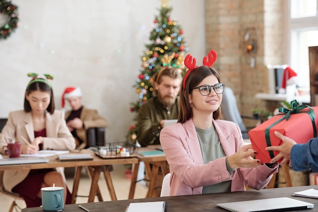 Gerente mujer morena joven feliz en diadema de Navidad tomando caja de regalo llena de manos de colega mientras está sentado en el lugar de trabajo en la oficina