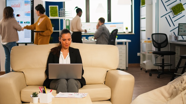 Gerente de mujer sosteniendo una computadora portátil, mirando en internet mientras está sentado en el sofá sonriendo debido a las buenas noticias. Compañeros de trabajo multiétnicos hablando de empresa financiera de inicio en la oficina moderna.