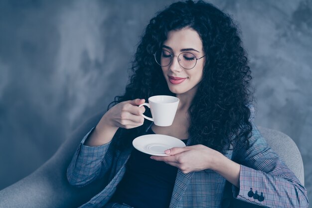 Gerente de niña de pelo ondulado sentarse en una silla bebiendo café caliente aislado sobre muro de hormigón