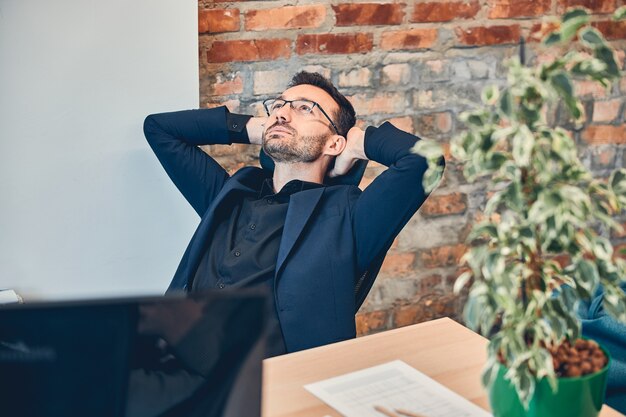 Gerente de proyecto exitoso sosteniendo la mano detrás de la cabeza y disfrutando el momento mientras está sentado en el lugar de trabajo moderno